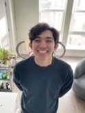 A young man with short brown hair wears a navy blue sweater and smiles at the camera with his hands behind his back in front of a large window and bicycle in an apartment.